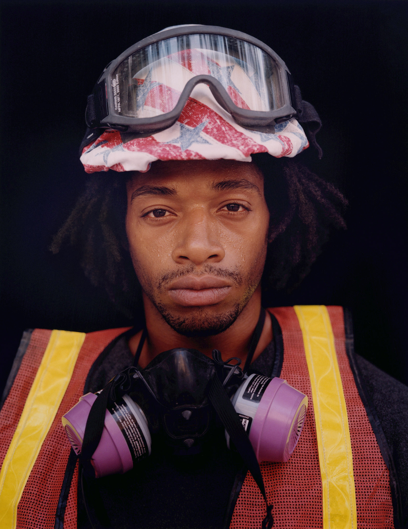 A man with goggles, safety vest, hard hat, adorned with stars and stripes, stares straight ahead in this photographic portrait.   