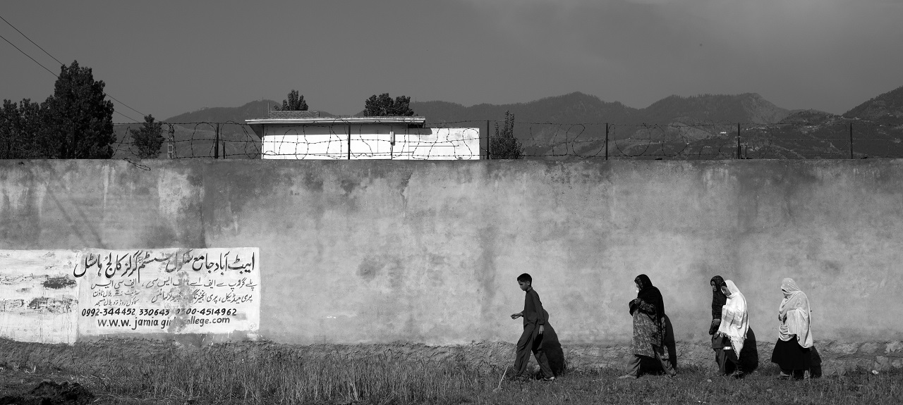 Four people walking along a long wall. A white building, mountaintops and sky are visible behind the wall. 