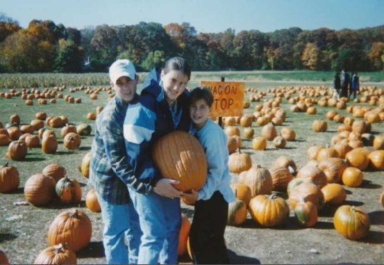 Three teenagers pose tightly hugging each other and holding a large pumpkin in a pumpkin patch. 