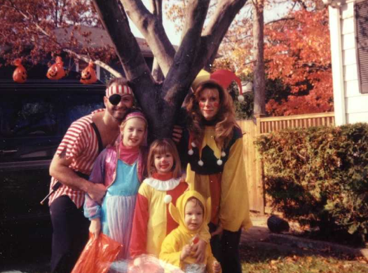A family fully costumed for Halloween stands in a front yard, in the background are trees with vibrantly orange leaves. 
