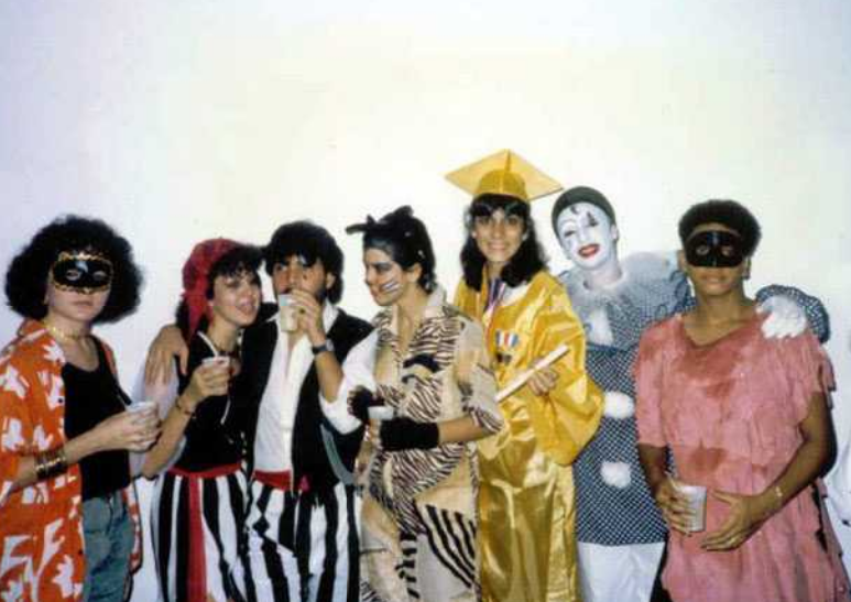 A group of teenagers stand in full Halloween costume in front of a white wall. 