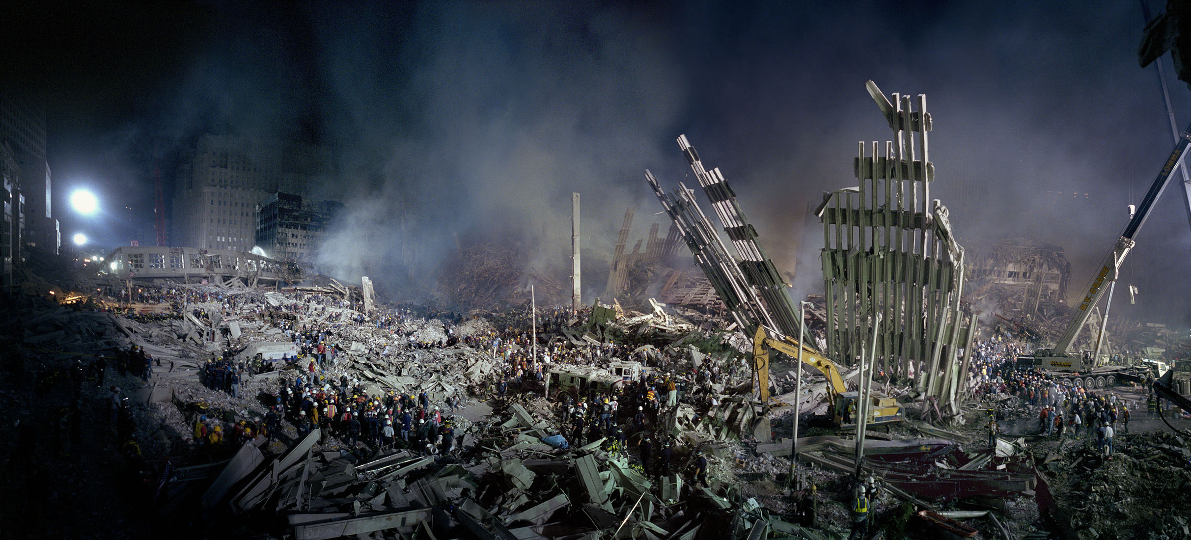 Wide view of Ground Zero ruins at night, with many workers in hardhats spread across the area. 