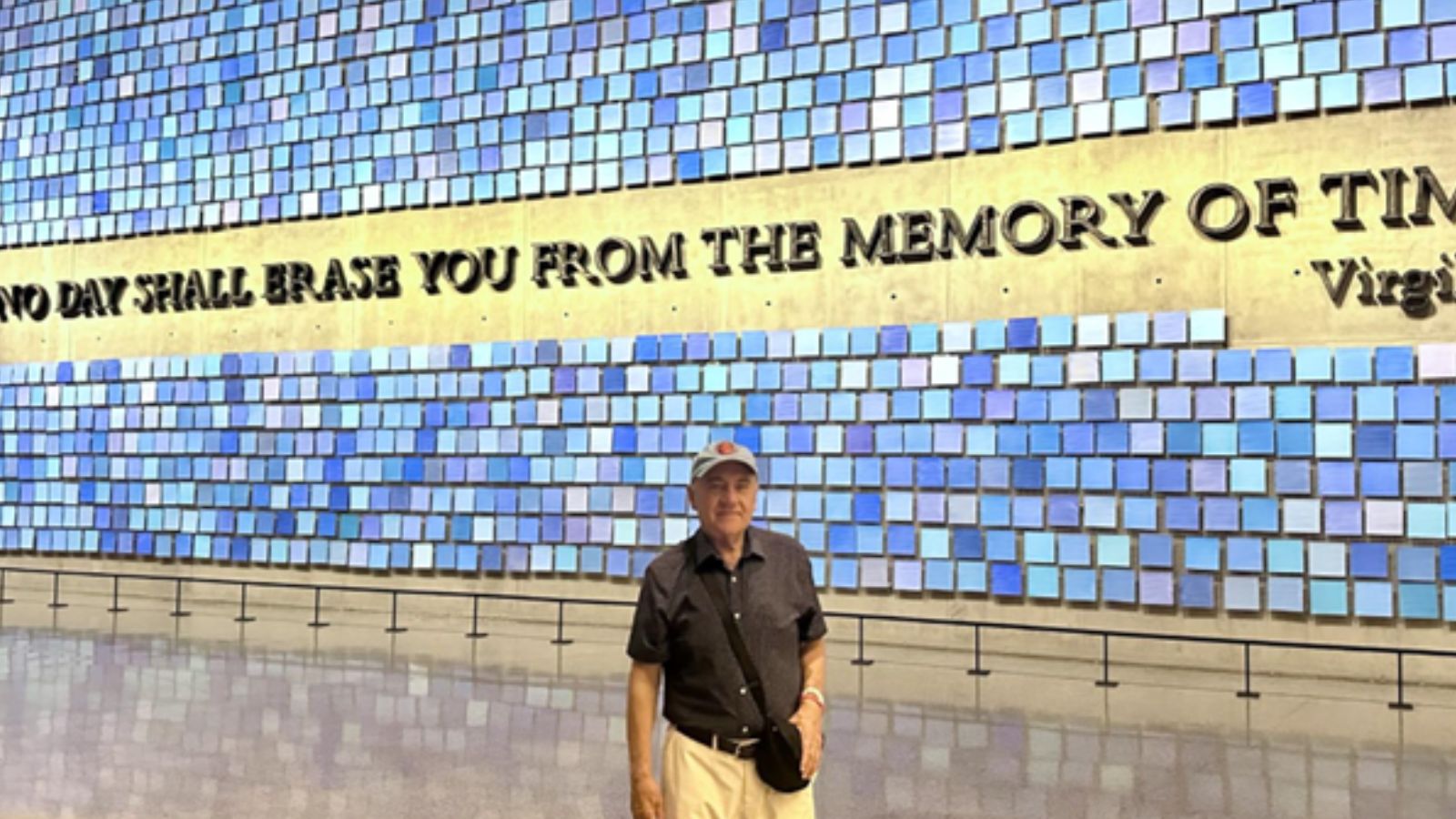 Frederick Agnostakis, in a black shirt and baseball cap, stands in front of a wall of square blue tiles surrounding the phrase "No Day Shall Erase You From the Memory of Time" by Virgil