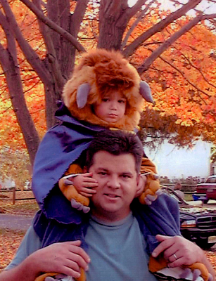 A man holds his young son, dressed as Beast, on his shoulders. In the background are trees with vibrantly orange leaves.