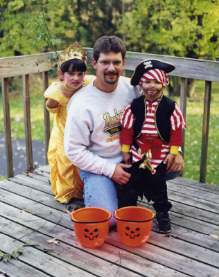 A man kneels on a deck beside his daughter, dressed as a princess, and his son, dressed as a pirate, on Halloween.