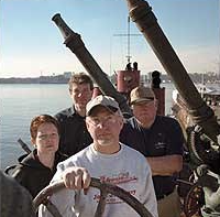 A group of men and woman at the captain's wheel of a small boat, with river  in the background