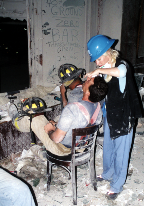 A woman with blonde hair in a blue hard hat tends to a man in a chair with his head bent back, amid a backdrop of rubble