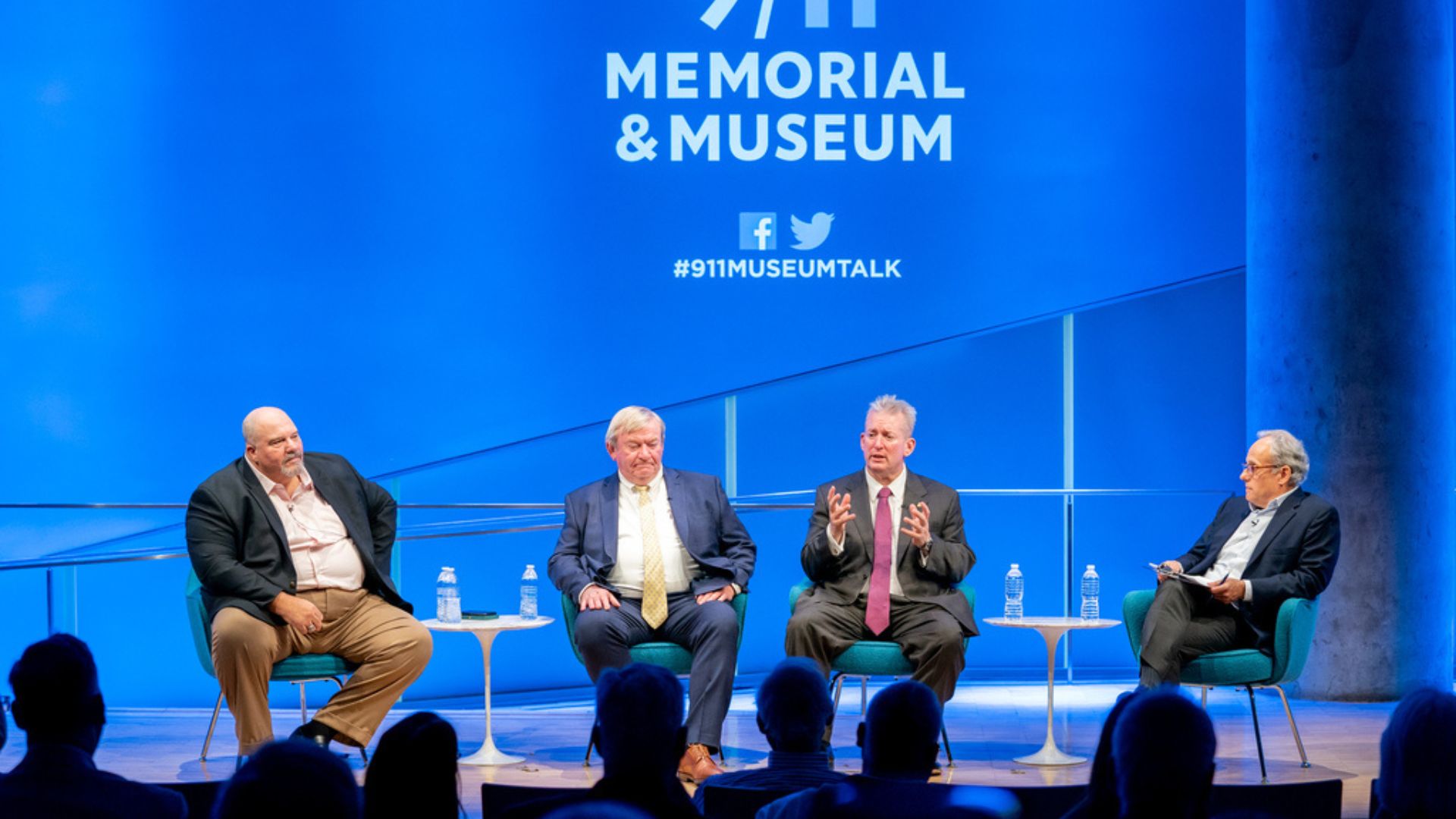 Four men on stage against a bright blue backdrop with the 9/11 Memorial & Museum logo