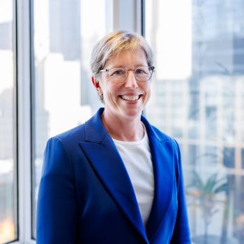 Beth Hillman, the President and CEO of the 9/11 Memorial and Museum, stands in front of a window with part of the New York City skyline behind her. She wears a bright blue blazer and a white shirt under it.