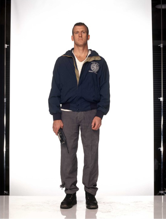 Tim Brown, dressed casually, stands in front of a white backdrop holding a walkie talkie at his side