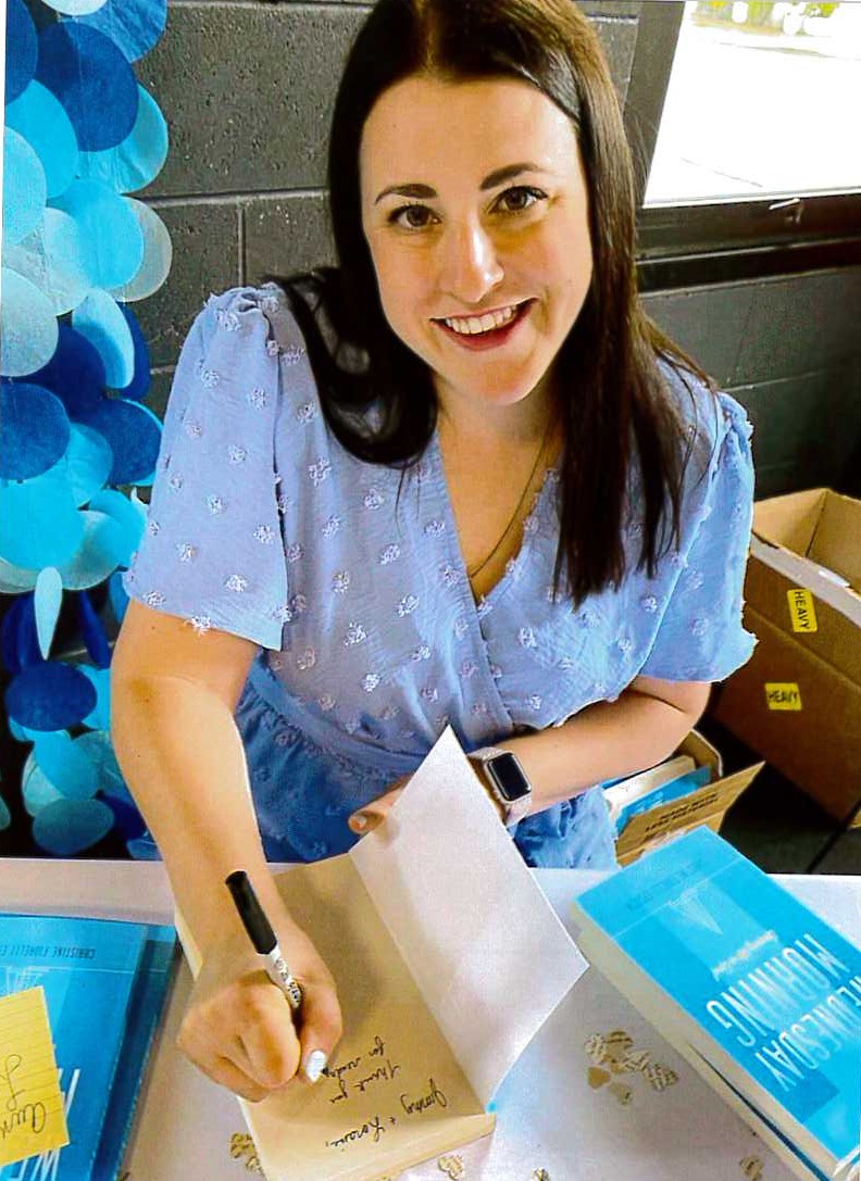 A woman with long straight hair in a blue floral dress smiles as she signs a book.