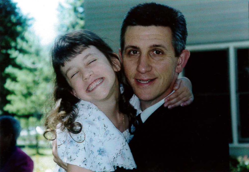 A young girl embraces her father, smiling so broadly that her eyes are closed.