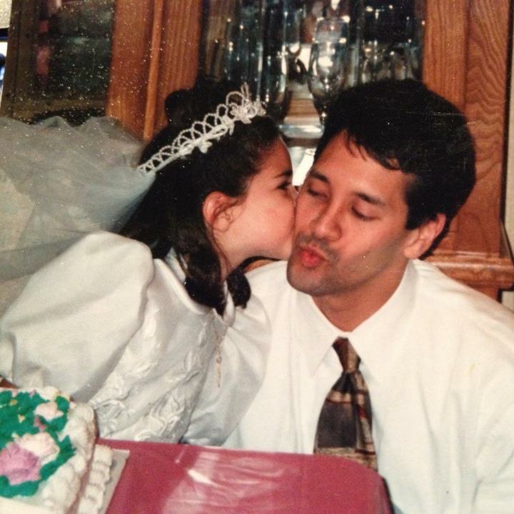 A girl in a white dress and bridal headband kisses her father on the cheek.