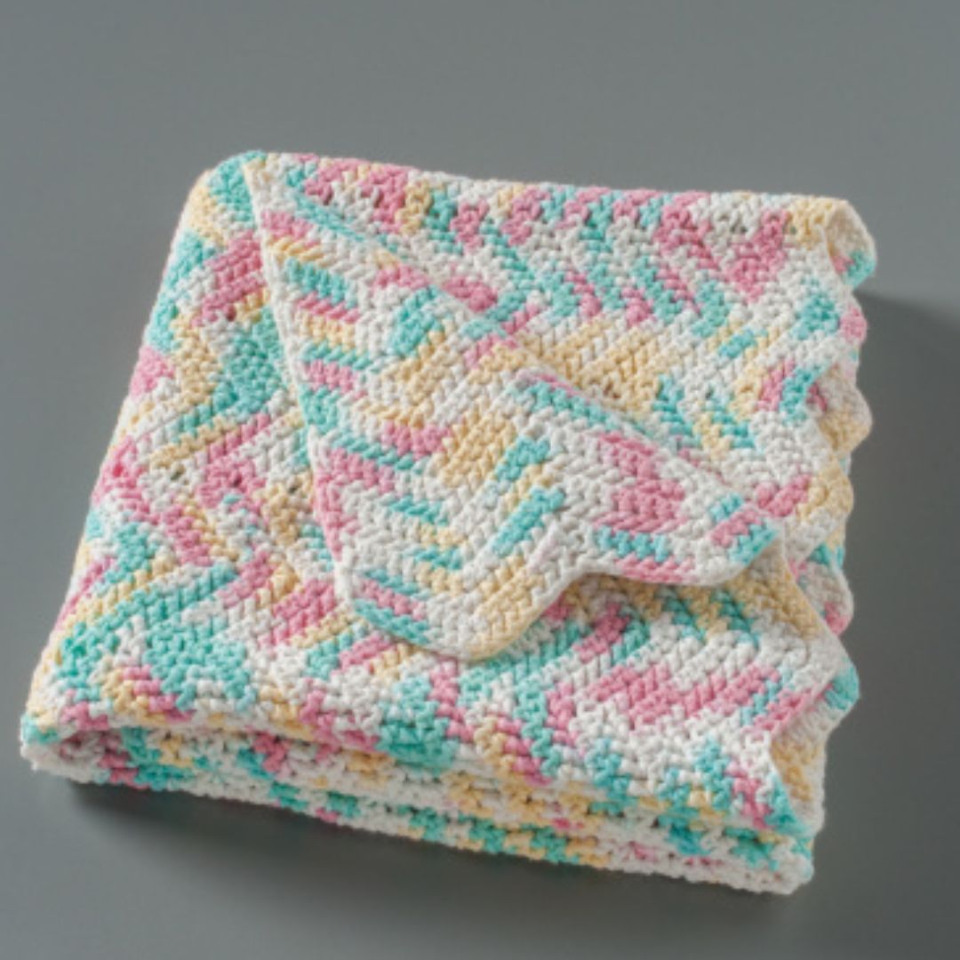 A crocheted blanket with scalloped edges, folded into a square with one corner dog-eared. The stitching is mint green, dark pink, pale yellow, and white. It is set against a gray background.