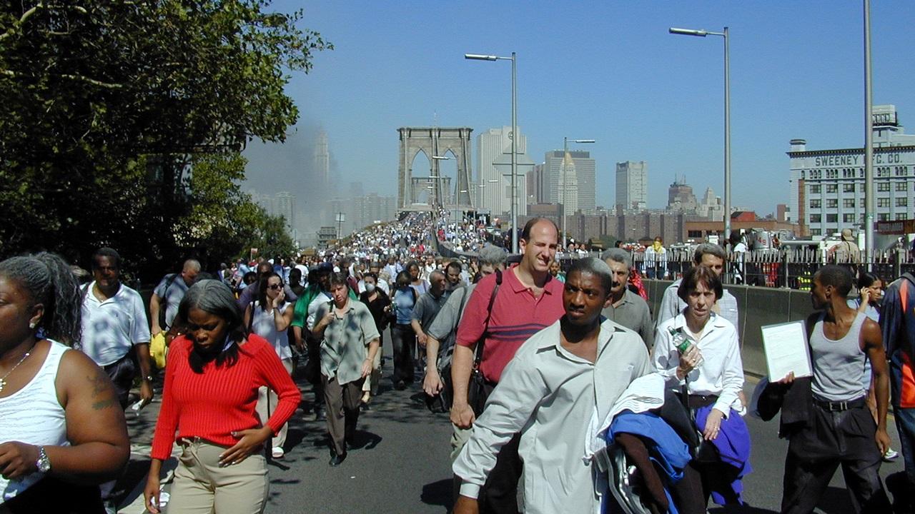 Dozens of people approach after walking across the Brooklyn Bridge on September Eleventh. They have dust and soot from the World Trade Center in their hair. Behind them, hundreds more people cross the bridge. In the distance is the skyline of lower Manhattan, with a cloud of smoke hovering over downtown.