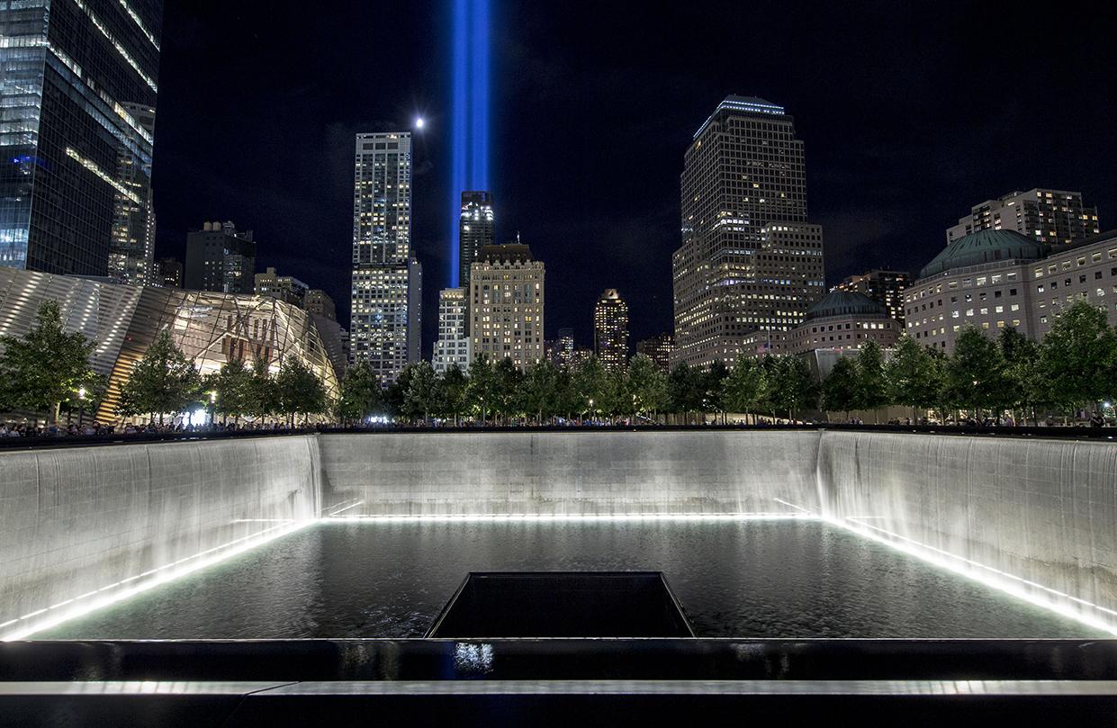 Water cascades down the illuminated walls of the North Tower reflecting pool on a warm night. The water pours down a square hole at the center of the pool. In the distance, a moon hangs over the city and the Tribute in Light shines above the buildings.