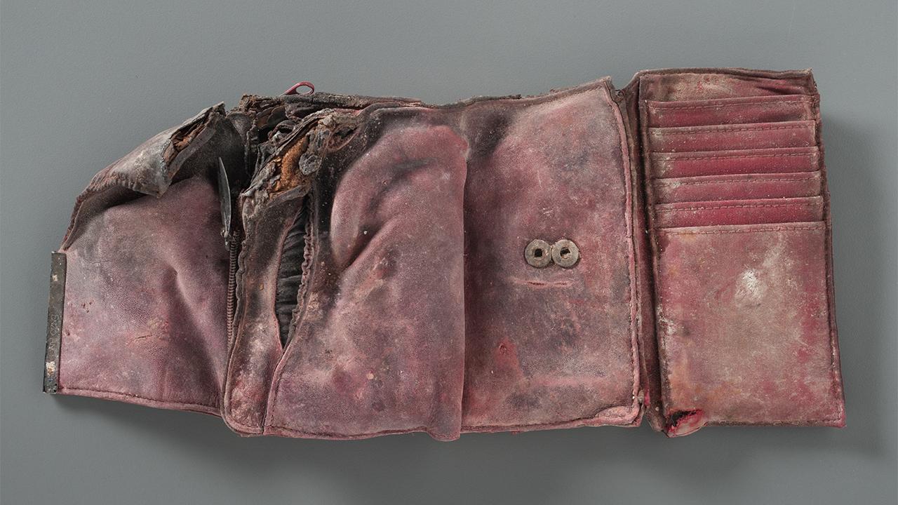 A damaged and stained red wallet from the Collection of the 9/11 Memorial Museum is open on a gray table. The wallet is empty. Its left corner is burned and ripped