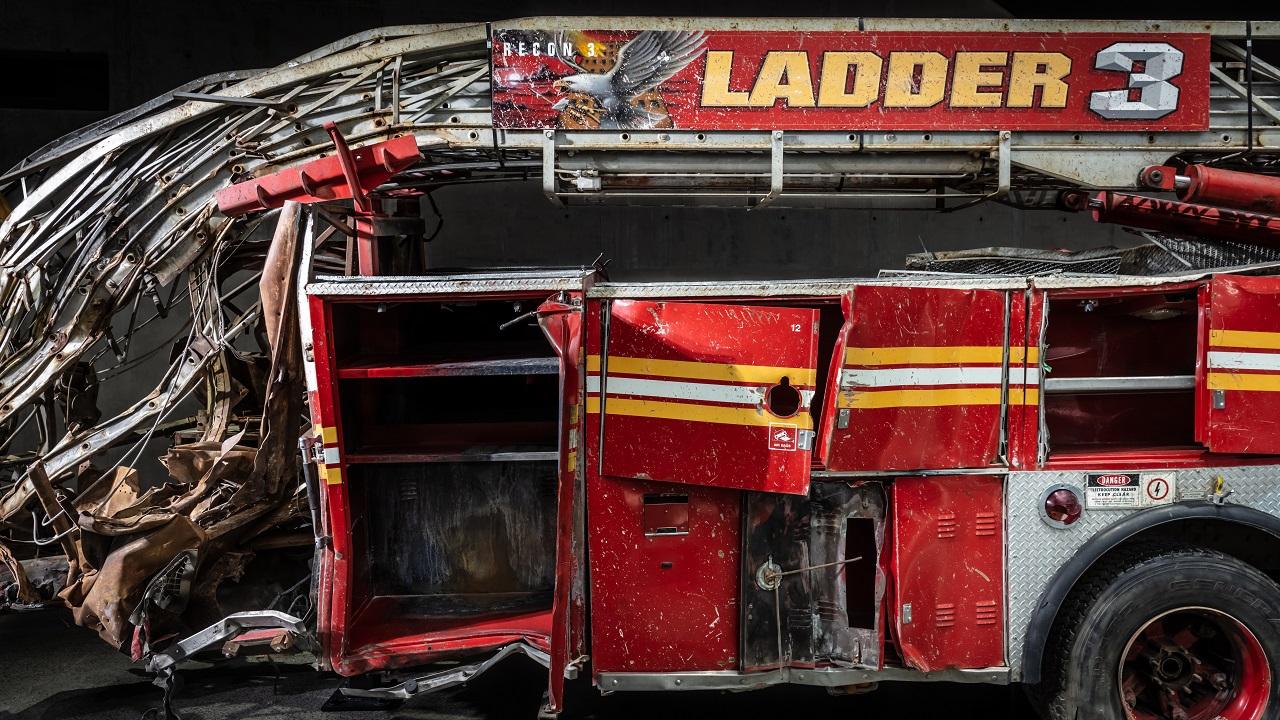 The heavily damaged firetruck of FDNY Ladder Company Three sits in the Museum. This close-up view shows the bright red vehicle’s twisted ladder and broken compartment doors.