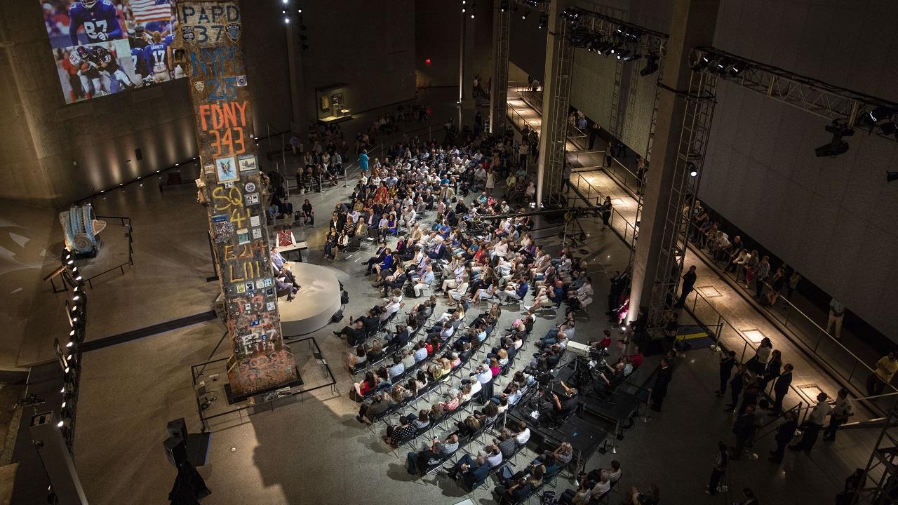 Dozens of seated people watch a video projected on a wall of Foundation Hall. The people are sitting by the base of the Last Column. The lighting in the hall has been dimmed for the event. Dozens more people stand and lean against railings off to the right as they also watch the video.