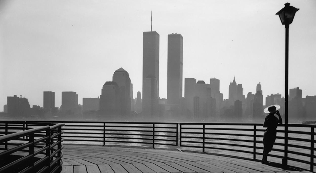 A woman in silhouette leans against a lamppost on a curved boardwalk, facing the view of lower Manhattan in the background. The Twin Towers, at the center of the skyline, rise into the cloudy sky.