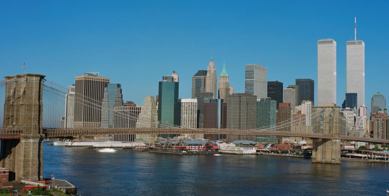 View of lower Manhattan's buildings behind a bridge spanning a calm river. The Twin Towers stand tall above other buildings, reaching up to a clear blue sky.