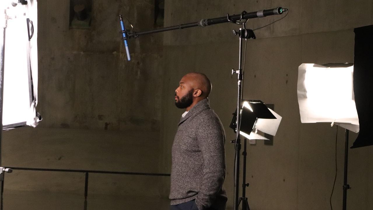 In this photograph a man stands in profile before a boom mic and set lights during the Anniversary in the Schools webinar filming