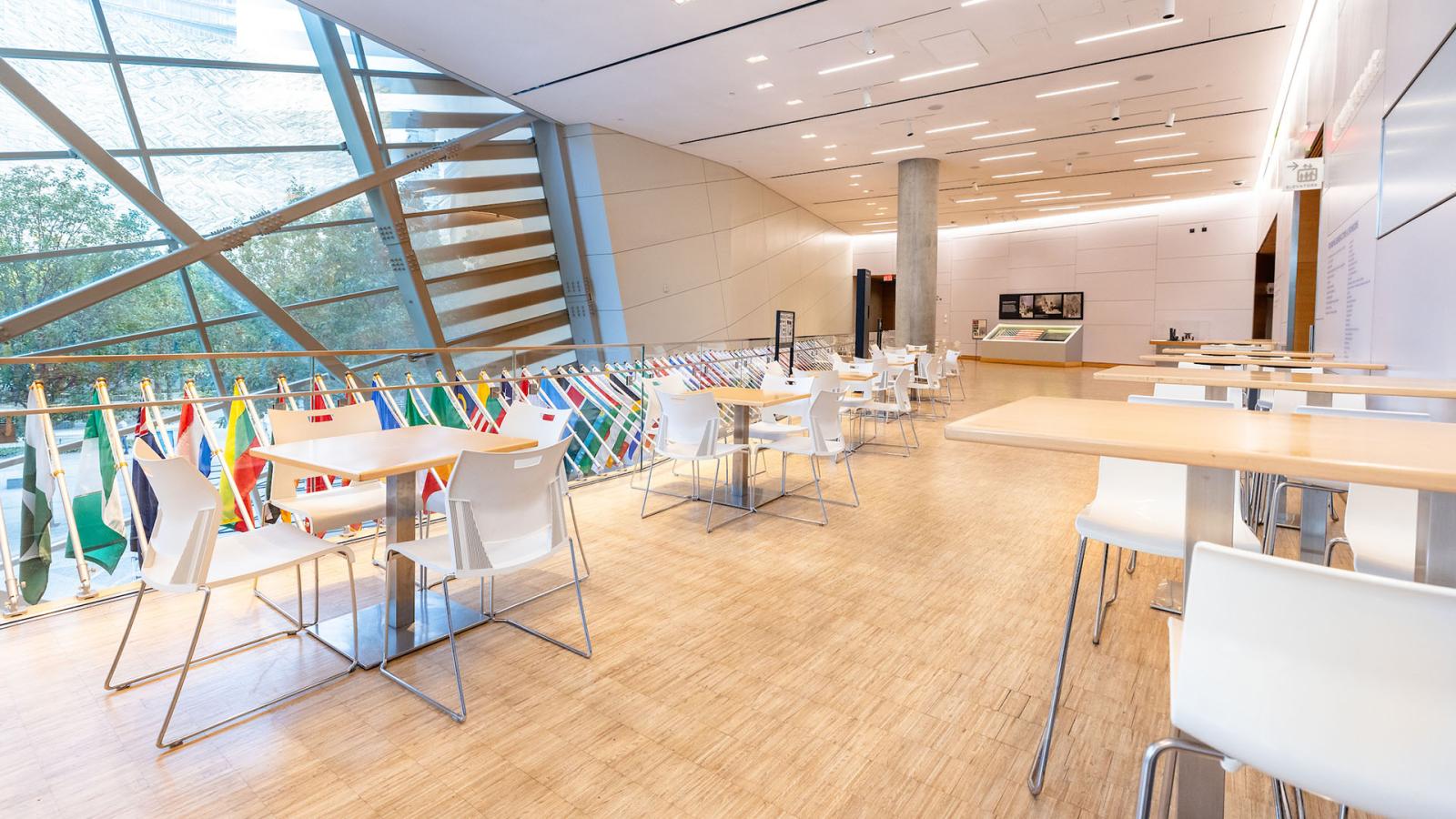 Cafe tables and chairs arranged on an atrium across from a large window and row of flags.