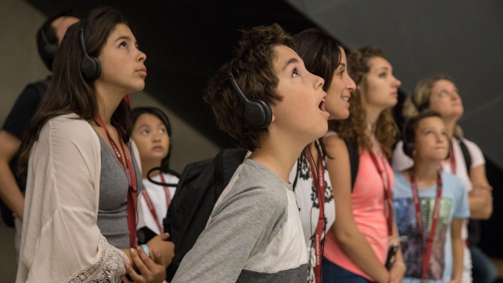 A group of young people, some wearing headsets, look up at an artifact in the Museum