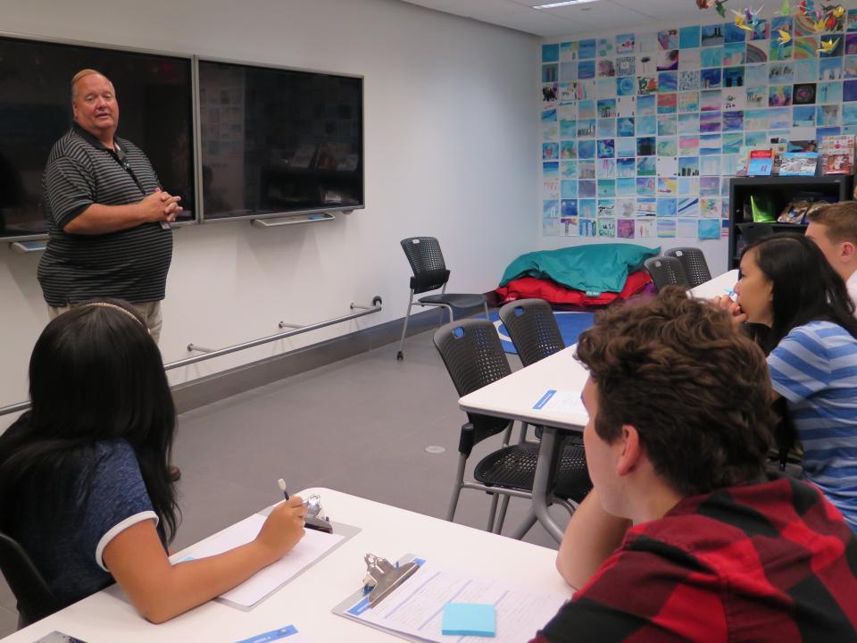 Firefighter Bill Spade stands in front of a smartboard as he speaks to young people at the Museum’s Education Center. The young people are seated at tables with clipboards.