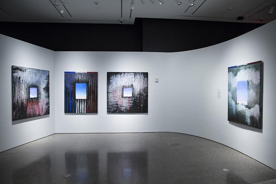 Four of Ejay Weiss’ paintings are seen on display at the 9/11 Memorial Museim. They are a part of his 9/11 Elegies series. The paintings are on square-shaped canvases. Square-shaped holes painted the color of the sky are at the center. These squares are surrounded by various abstract designs in black, white, red, and blue.