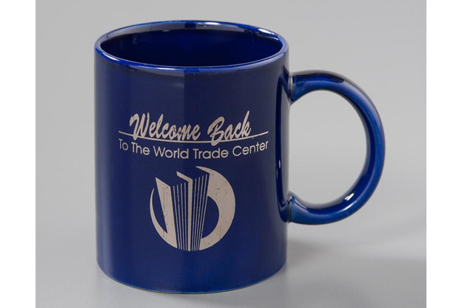 A blue “Welcome Back” mug is displayed on a white surface at the Museum. The mug features a depiction of the Twin Towers with the words, “Welcome Back to the World Trade Center.”