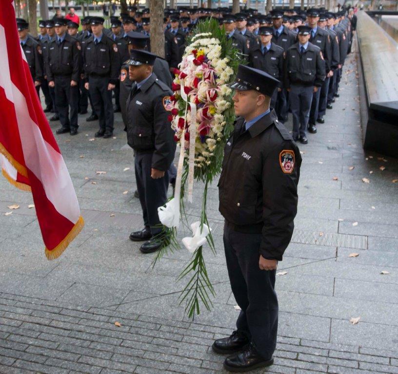 FDNY firefighter Brendan Stackpole stands beside a wreath on 9/11 Memorial plaza during an FDNY tribute. Dozens of other FDNY firefighters stand in the distance.