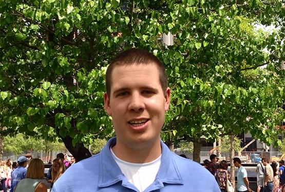 Tom Brown smiles for a photo in front of the Survivor Tree at Memorial plaza on a sunny day.