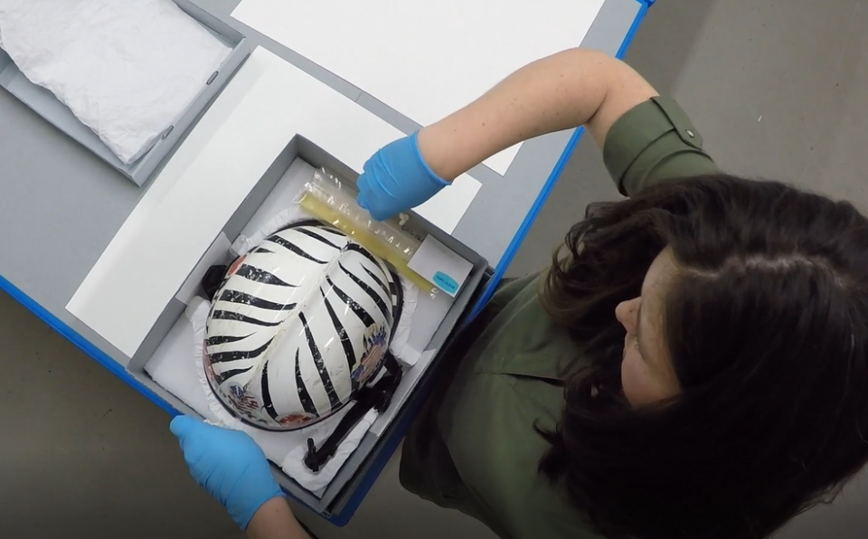 A Museum employee wears blue gloves as she removes a rescue and recovery helmet from a box.