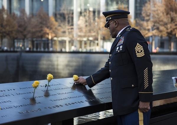 A man in a formal military outfit places a yellow rose at a victim’s name on a bronze parapet at the 9/11 Memorial. Two other yellow roses have been placed at names nearby.