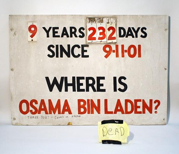 A sign that tracked the days Osama bin Laden was at large is displayed on a white surface. The sign reads: “9 years, 232 days since 9/11/01. Where is Osama bin Laden?” A yellow piece of paper beside the sign reads “Dead.”