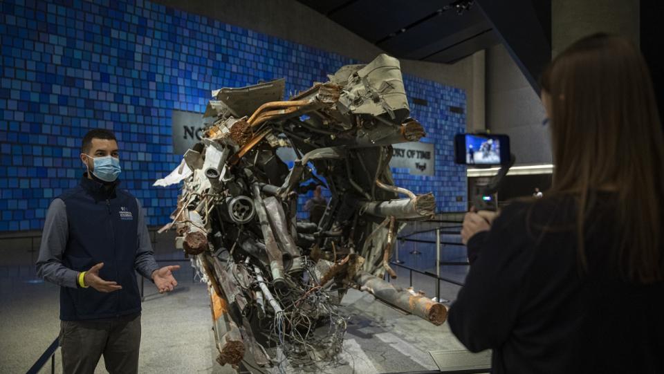 A tour guide wearing a mask stands beside a piece of the mangled antenna. A woman films him speaking on a smartphone.