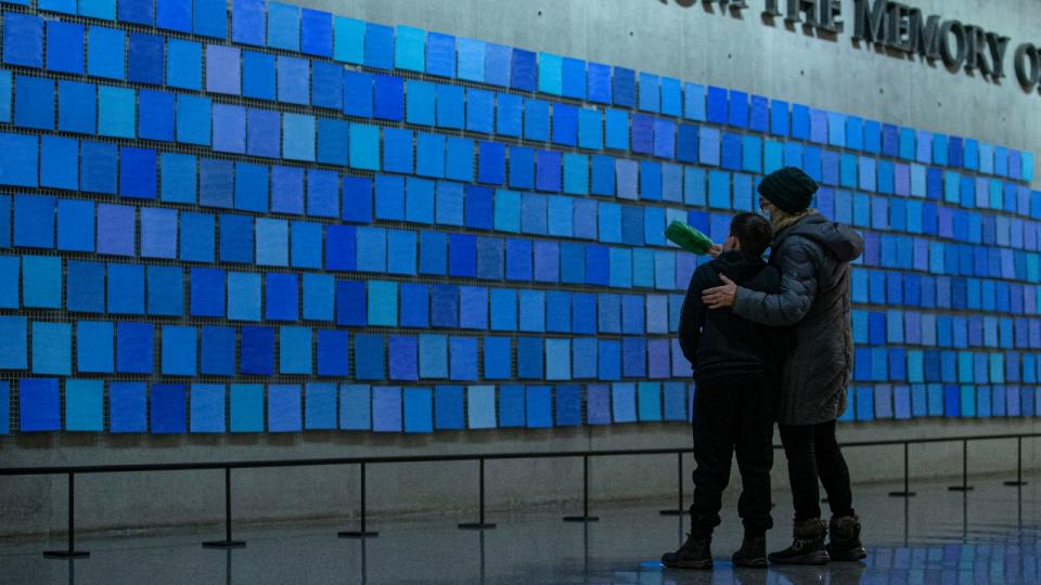 An older woman wraps her arm around a young boy's shoulder as they look at the inscription "No Day Shall Erase You From the Memory of Time.” This quote from Book IX of "The Aeneid" by the Roman poet Virgil are are part of a larger art installation by artist Spencer Finch, titled “Trying to Remember the Color of the Sky on That September Morning.” The words appear amid a sea of blue tiles.