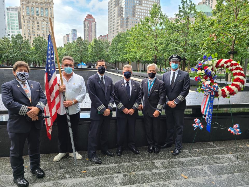 Five men in pilot uniforms along with another in white top and blue pants stand at the Memorial with an American flag and a commemorative wreath